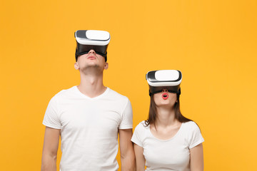Amazed young couple friends guy girl in white empty blank t-shirts posing isolated on yellow orange background. People lifestyle concept. Mock up copy space. Watching in headset of vr virtual reality.