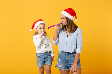 Fototapeta premium Woman child baby girl in Christmas Santa hat blow in pipe. Mommy little kid daughter isolated on yellow background studio portrait. Happy New Year 2020 celebration holiday concept. Mock up copy space.