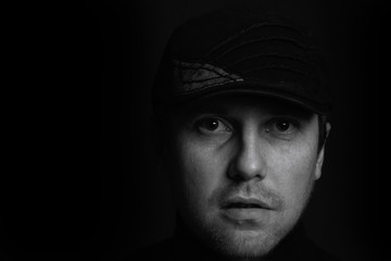 Black and white closeup portrait of an unshaven serious young gangster man with a cap on a dark background.