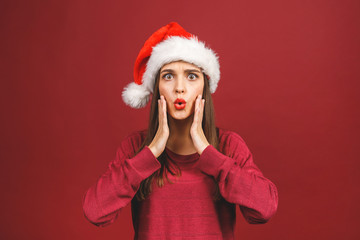 Attractive Woman With Santa Hat Looking Surprised And Amazed isolated against red background.