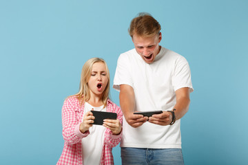 Young funny couple two friends guy girl in white pink blank t-shirts posing isolated on pastel blue background. People lifestyle concept. Mock up copy space. Playing video game with mobile phones.