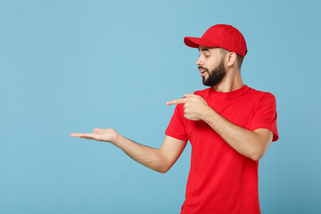 Delivery man in red uniform workwear isolated on blue wall background, studio portrait. Professional male employee in cap t-shirt print working as courier dealer. Service concept. Mock up copy space.