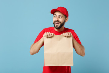 Delivery man in red uniform hold craft paper packet with food isolated on blue background, studio portrait. Male employee in cap t-shirt print working as courier. Service concept. Mock up copy space.