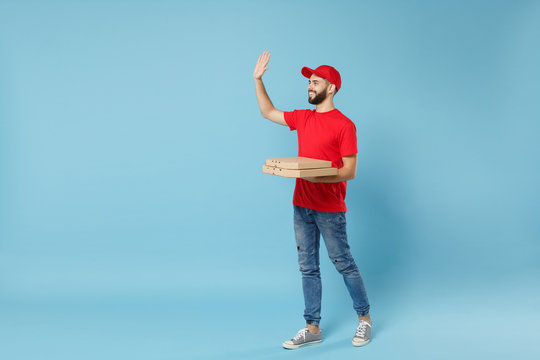 Delivery man in red workwear giving food order pizza boxes isolated on blue background, studio portrait. Professional male employee in cap t-shirt print courier. Service concept. Mock up copy space.
