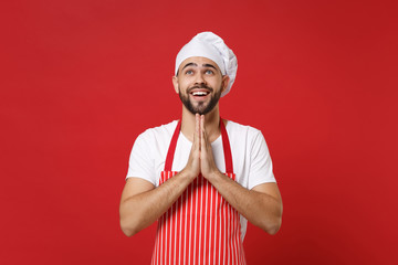 Smiling bearded male chef cook or baker man in striped apron white t-shirt toque chefs hat posing isolated on red background. Cooking food concept. Mock up copy space. Holding hands folded in prayer.