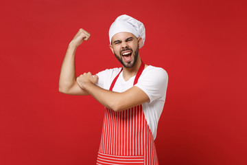 Strong young bearded male chef cook or baker man in striped apron white t-shirt toque chefs hat posing isolated on red background. Cooking food concept. Mock up copy space. Showing biceps, muscles.