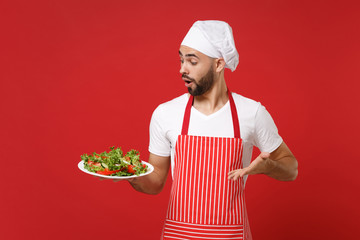 Shocked young bearded male chef cook or baker man in striped apron toque chefs hat posing isolated on red wall background. Cooking food concept. Mock up copy space. Hold plate with vegetable salad.