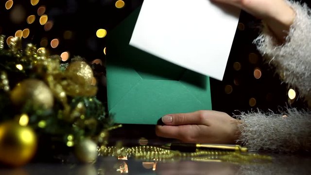 Female hands are opening green envelope with blank sheet of paper for greeting card. Decorated wreath, decor for christmas tree, balls are on table. Garland with yellow light bulbs are blinking. 