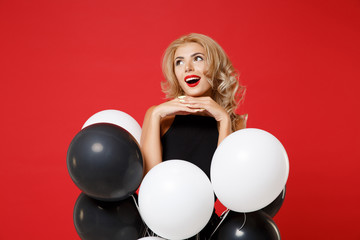 Pensive young woman girl in black clothes posing isolated on red background. Women's Day birthday holiday party concept. Mock up copy space. Celebrating holding air balloons put hands prop up on chin.