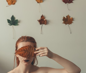 Red-haired girl with dark lips closes her eyes with a yellow dried leaf on the white background with red green yellow maple leaves on it in three rows of three -autumn theme