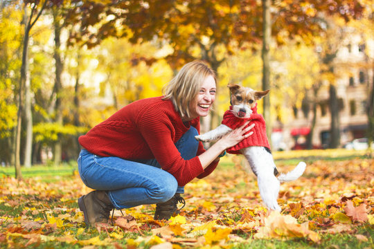 Stylish hipster woman playing with her puppy Jack Russell in an autumn park, autumn mood. Romantic day with a pet. The dog wears a sweater. Happy smiling girl looking directly at the camera