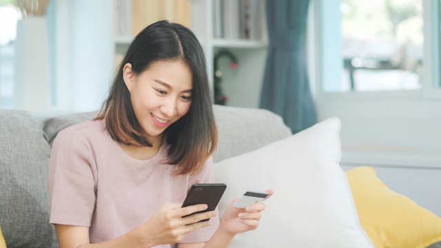 Young smiling Asian woman using smartphone buying online shopping by credit card while lying on sofa when relax in living room at home. Lifestyle latin and hispanic ethnicity women at house concept.