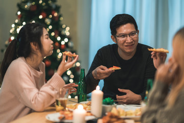 Merry Christmas and Happy New Year! Young friends group having dinner at home. Asian family party with beer and pizza food near christmas tree at house in night. Celebration holiday and togetherness.