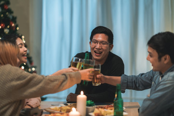 Merry Christmas and Happy New Year! Young friends group having dinner at home. Asian family party with beer and pizza food near christmas tree at house in night. Celebration holiday and togetherness.