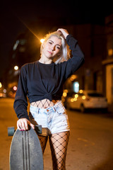 Front view of young pretty woman looking camera in shorts while standing on the street holding a skateboard at night in the city