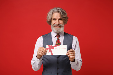 Smiling elderly gray-haired mustache bearded man in classic shirt vest and colorful tie isolated on red background in studio. People lifestyle concept. Mock up copy space. Holding gift certificate.