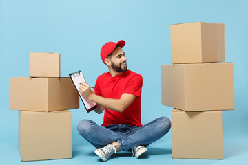 Delivery man in red uniform isolated on blue background, studio portrait. Male employee in cap t-shirt print working as courier dealer sit at empty cardboard box. Service concept. Mock up copy space.