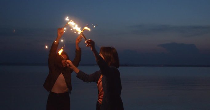 Two young women with fireworks celebrate, dance, spin. In the evening near the sea, against the background of blue sky and moon.