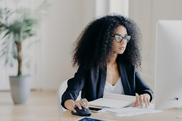 Serious curly businesswoman focused at display of computer, works on making project, surrounded...