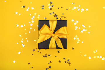 Black gift box with yellow bow on yellow background with sparkling confetti. Flat lay.