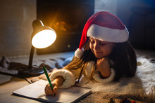 Little girl in Santa hat writes letter to Santa Claus near christmas tree and clock