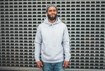 City portrait of handsome hipster guy with beard wearing gray blank hoodie or sweatshirt and hat...
