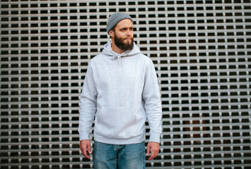 City portrait of handsome hipster guy with beard wearing gray blank hoodie or sweatshirt and hat...