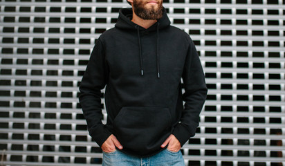 City portrait of handsome hipster guy with beard wearing black blank hoodie or sweatshirt and hat with space for your logo or design. Mockup for print