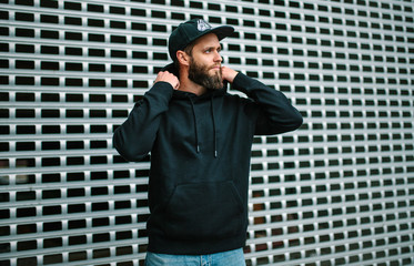 City portrait of handsome hipster guy with beard wearing black blank hoodie or sweatshirt and hat...
