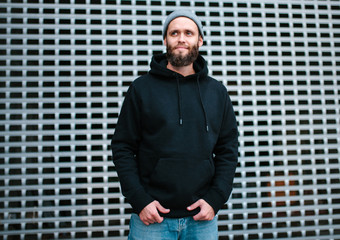 City portrait of handsome hipster guy with beard wearing black blank hoodie or sweatshirt and hat...