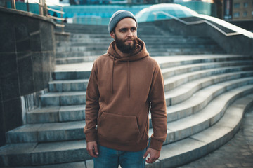 City portrait of handsome hipster guy with beard wearing brown blank hoodie or hoody and hat with...