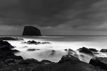 Seascape on a cloudy early evening with vulcaninc rocks on foreground and a large sea rock in the background, Black and White Photo, Madeira Island, Portugal