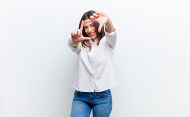 young pretty woman feeling happy, friendly and positive, smiling and making a portrait or photo frame with hands isolated against white wall