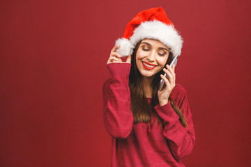 Young girl in a Santa Claus hat and with mobile phone in hand isolated on a red background.