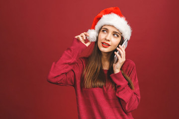Young girl in a Santa Claus hat and with mobile phone in hand isolated on a red background.