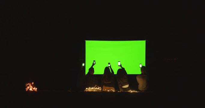 People take pictures of the movie theater screen on a mobile phone. Friends watch a movie in an open-air cinema. At night on the beach near the fire. Green screen