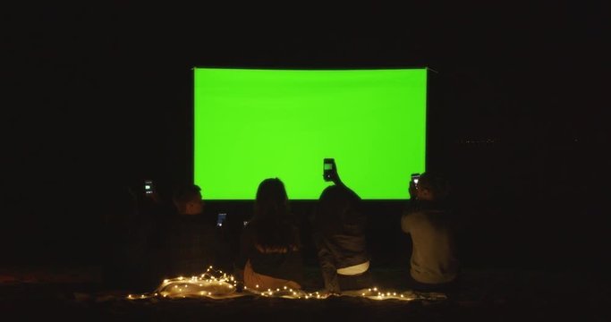Women and guys are resting and taking pictures of the screen of an open-air cinema. Green screen.