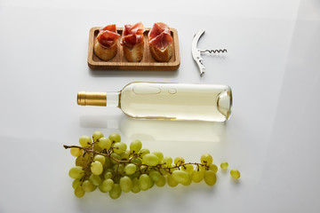 flat lay with bottle with white wine near grape, corkscrew and sliced prosciutto on baguette on white background