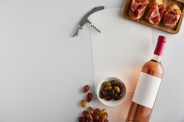 top view of bottle with rose wine near grape, corkscrew, olives, blank paper and sliced prosciutto on baguette on white background
