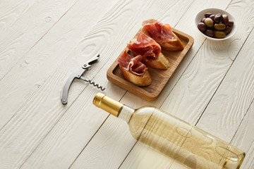 bottle with white wine near prosciutto on baguette, olives and corkscrew on white wooden surface