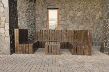 Patio on the background of a stone wall with wooden benches around a low wooden table outdoors against the background of windows in which the sky is reflected.
