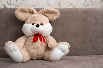 Fototapeta premium Plush rabbit sitting on a brown sofa. Beige toy rabbit with a red bow. Place for text. Romantic card
