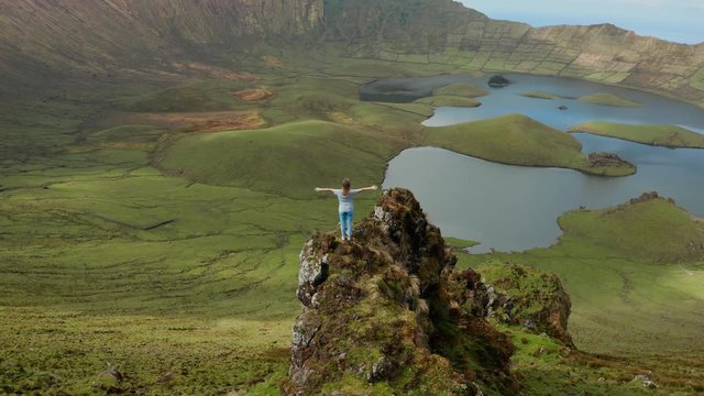 Girl with wide open arms on a rock above grate and beautiful green caldera of inactive volcano. Aerial of Corvo, Azores