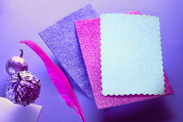 Creative purple and pink Halloween background with levitating pink pin quill, stack of glittering paper and decorative pumpkins painted metallic pink, copy-space.