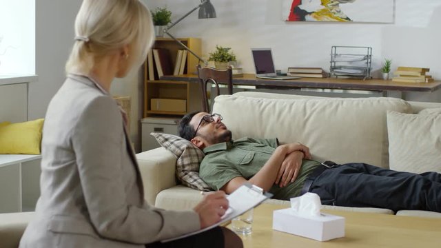 PAN of female psychotherapist nodding head and taking notes while listening to young middle eastern male patient lying on sofa and telling about problems during therapy session