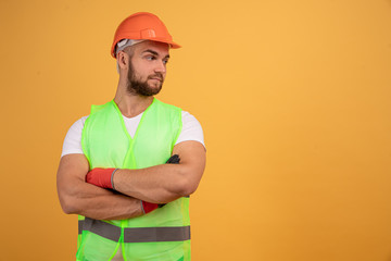 Shot of a pensive dissatisfied construction worker in protective orange helmet, stands with arms crossed, thinks over new building project, models over yellow wall. free space for text or logo