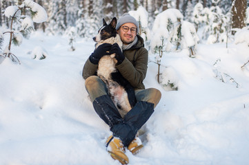 Fototapeta premium Happy man holding lovely dog in his hands in snowy forest. Smiling boy hugging adorable puppy in winter wood. Pet lover. Dog - human`s friend concept.