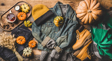 Autumn trendy outfit layout. Flat-lay of vintage denim jacket, sweater, jeans, woolen scarf, hat, yellow boots, pumpkins, candle and tea in cup over wooden background, top view. Fall apparel and mood