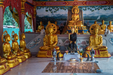 Obraz premium Bang Saen, Thailand - March 16, 2019: Wang Saensuk Buddhist Monastery. Golden statues in main open Prayer Hall with in center statue of founder of abbey. Green foliage outside.
