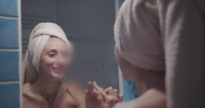 The Young Adult Woman Wipe Mirror Glass In Foggy Bathroom. The Girl Stay Wrapped Into White Towel And Smiling On The Mirorr.